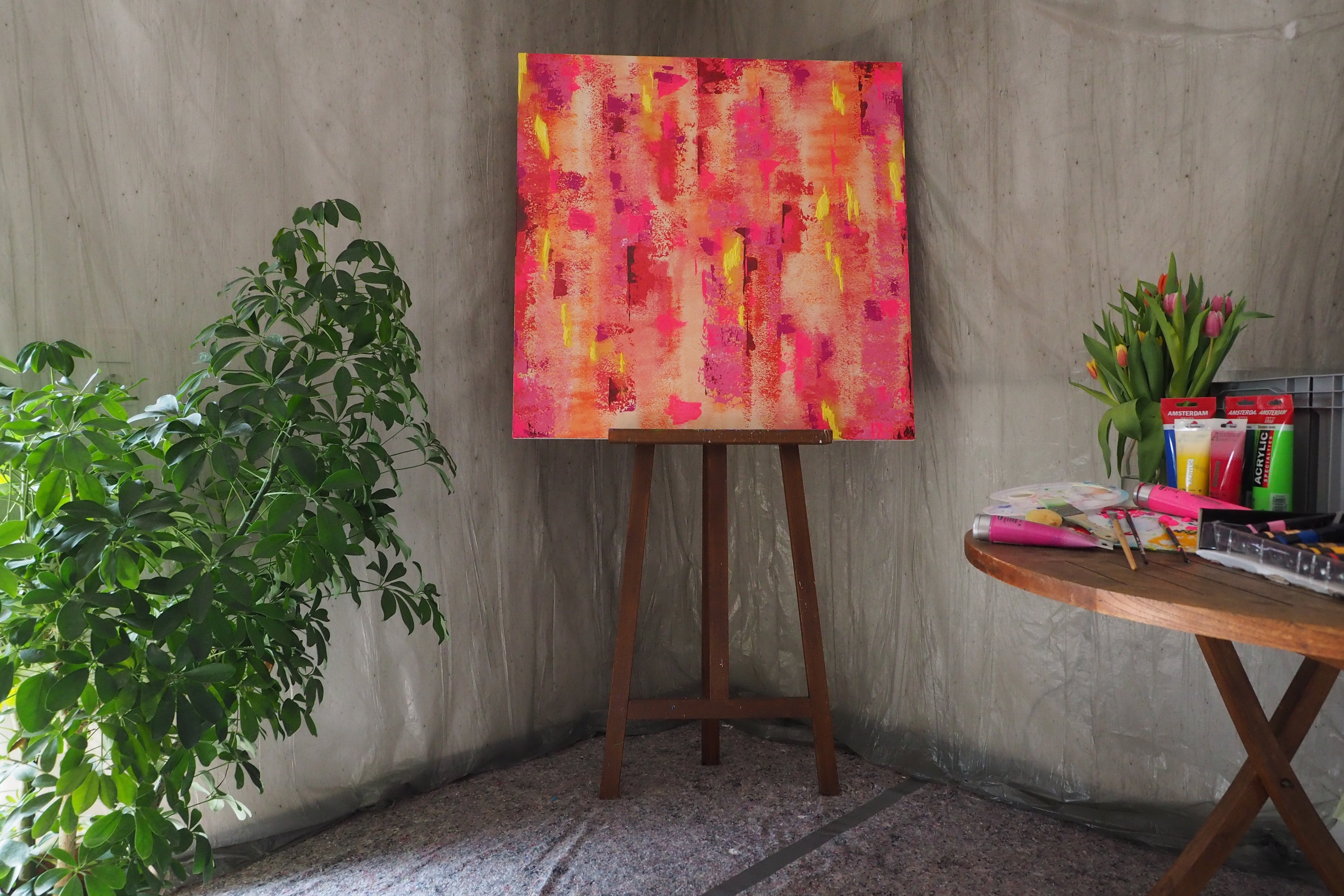 Das originale Kunstwerk „Lebensbaum“ von maridy art – ein leuchtendes abstraktes rosa und gelbes Gemälde, das Wachstum symbolisiert – steht auf einer Staffelei zwischen einer grünen Blattpflanze und einem runden Tisch mit Tulpen, Farbtuben und Künstlerbedarf in einem sanft beleuchteten Studio.
