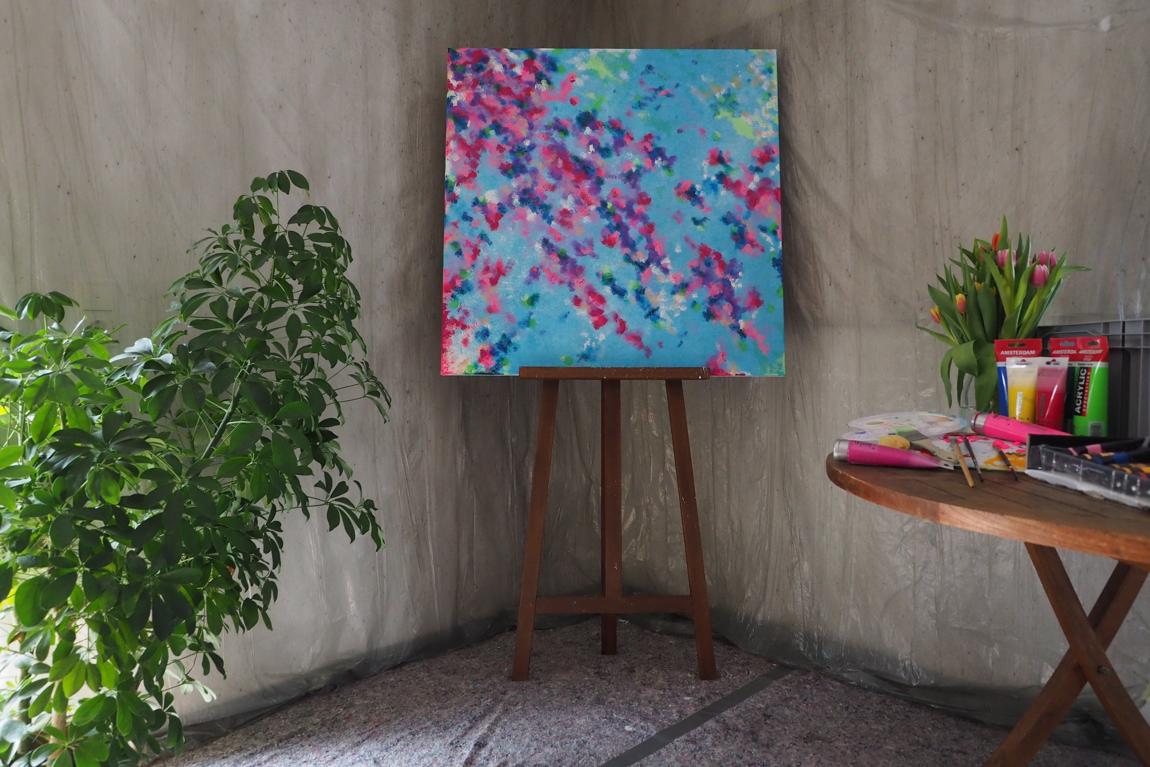 Das originale Kunstwerk „Blütenmeer“ von Maridy Art steht auf einer Staffelei vor einer grauen Wand neben einer grünen Pflanze. Ein runder Tisch mit Tulpen, Künstlerbedarf und Notizbüchern bringt Frische ins Ambiente.