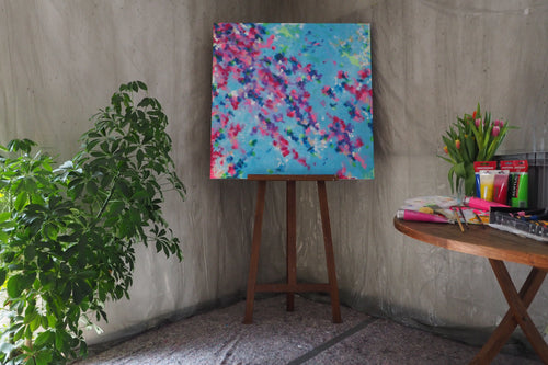 Das originale Kunstwerk „Blütenmeer“ von Maridy Art steht auf einer Staffelei vor einer grauen Wand neben einer grünen Pflanze. Ein runder Tisch mit Tulpen, Künstlerbedarf und Notizbüchern bringt Frische ins Ambiente.