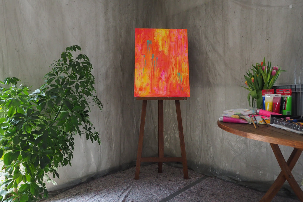 Das Originalkunstwerk „Feuerwerk“ von Maridy Art leuchtet farbenfroh auf einer Staffelei vor einer grauen Wand. Links steht eine grüne Pflanze, daneben ein runder Tisch mit Tulpen, Malutensilien und Pinseln für kreative Atmosphäre.