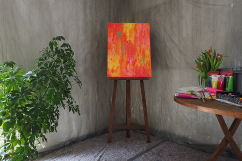 Das Originalkunstwerk „Feuerwerk“ von Maridy Art leuchtet farbenfroh auf einer Staffelei vor einer grauen Wand. Links steht eine grüne Pflanze, daneben ein runder Tisch mit Tulpen, Malutensilien und Pinseln für kreative Atmosphäre.