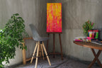 Das originale Kunstwerk „Sonnenstrahl“ von Maridy Art in Gelb und Rot steht auf einer Staffelei in einem gemütlichen Raum mit grauem Stuhl, grüner Pflanze und rundem Tisch mit Tulpen und Kunstbedarf, sanft von warmem Licht beleuchtet.