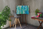 Ein grauer Stuhl steht neben einer Staffelei, auf der das Originale Kunstwerk „Mehr Meer“ von maridy art, ein abstraktes blau-grünes Gemälde, hängt. Eine Blattpflanze und ein Tisch mit Tulpen, Künstlerbedarf und Notizbüchern runden den gemütlichen, sanft beleuchteten Raum ab.