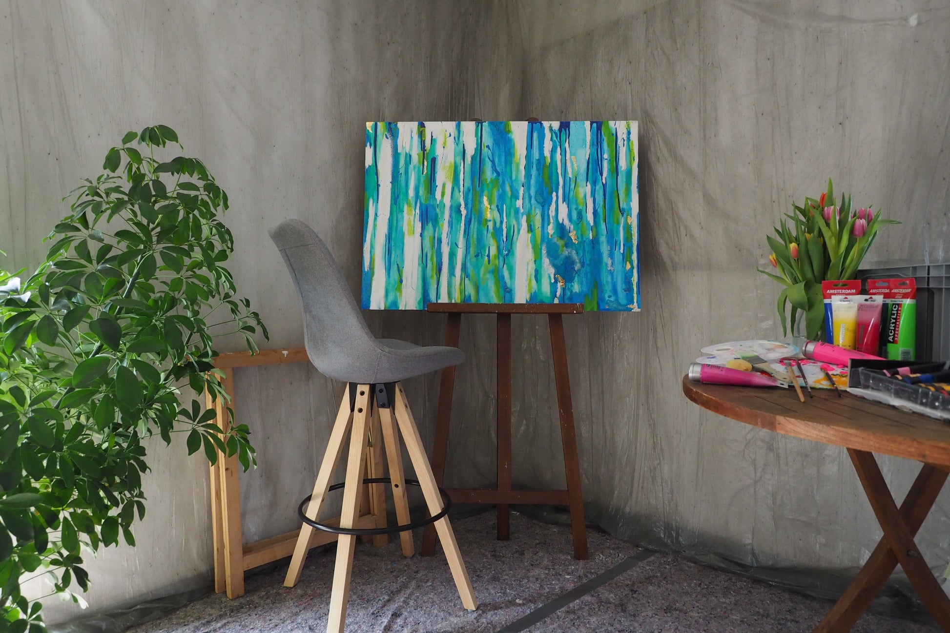 Ein grauer Stuhl steht neben einer Staffelei, auf der das Originale Kunstwerk „Mehr Meer“ von maridy art, ein abstraktes blau-grünes Gemälde, hängt. Eine Blattpflanze und ein Tisch mit Tulpen, Künstlerbedarf und Notizbüchern runden den gemütlichen, sanft beleuchteten Raum ab.
