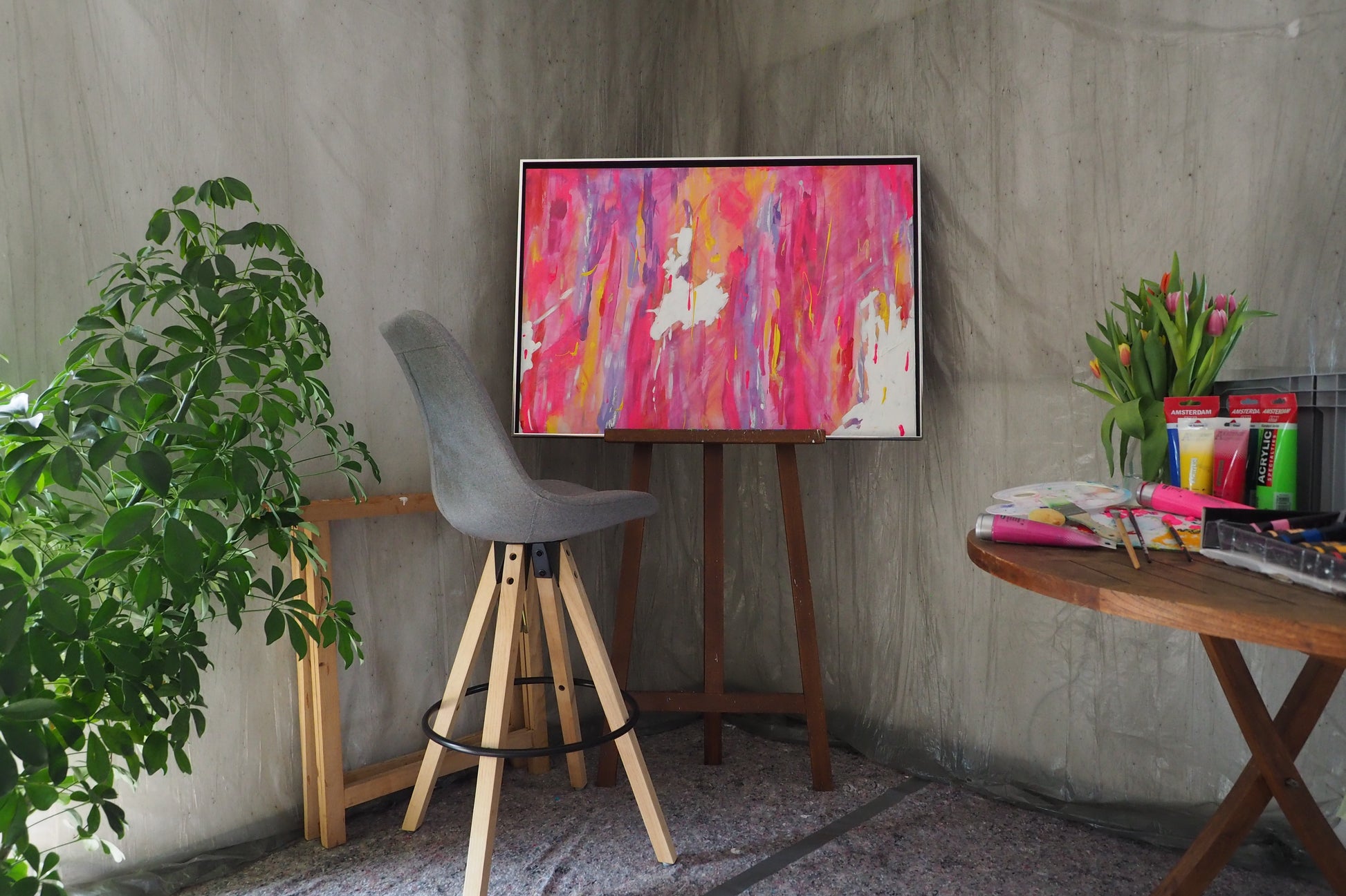Das originale Kunstwerk „Lebensfreude“ von Maridy Art steht auf einer Staffelei in einem Raum mit grauem Stuhl, grüner Pflanze, Tisch mit Kunstbedarf und Tulpen sowie strukturierter grauer Wand.