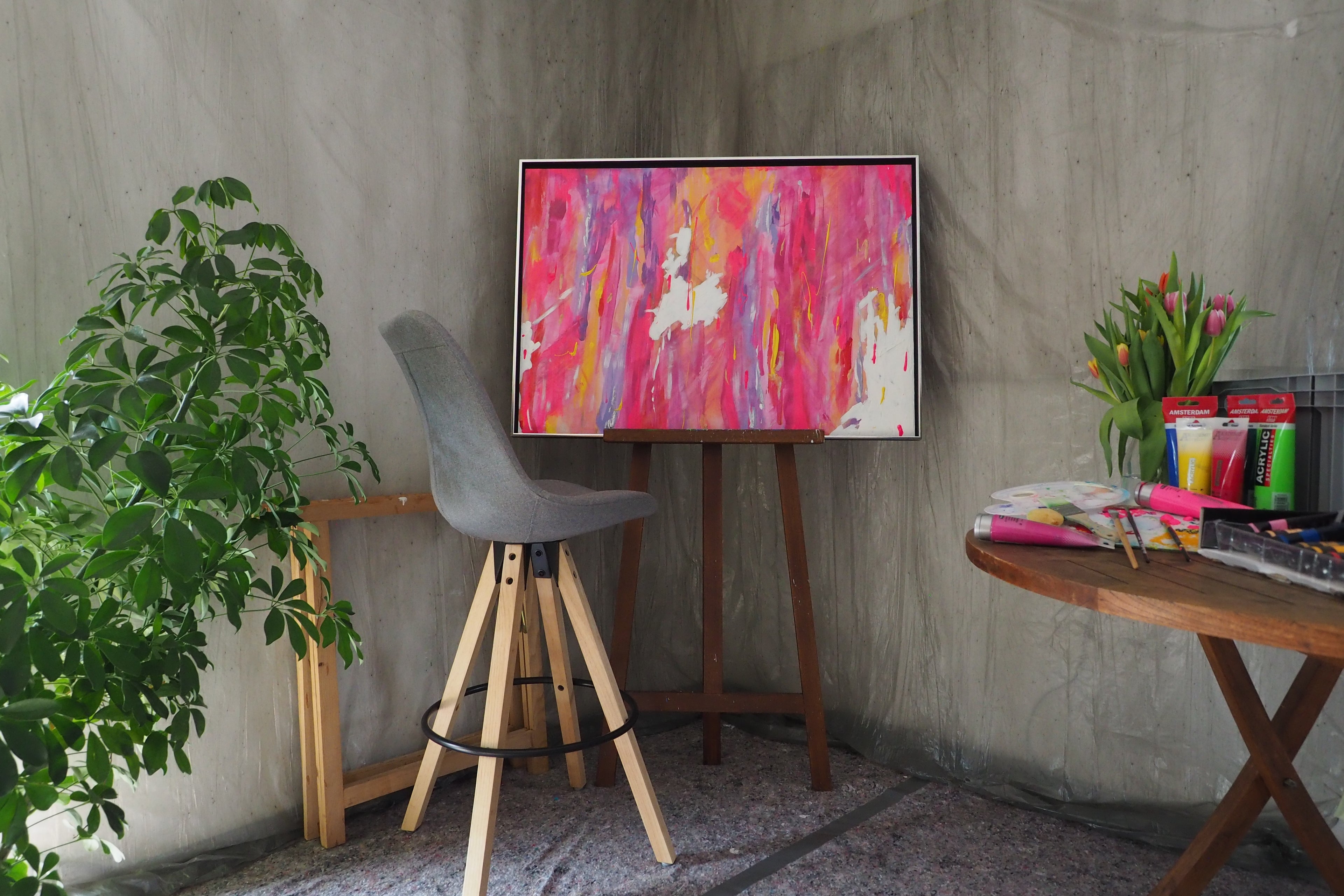 Das originale Kunstwerk „Lebensfreude“ von Maridy Art steht auf einer Staffelei in einem Raum mit grauem Stuhl, grüner Pflanze, Tisch mit Kunstbedarf und Tulpen sowie strukturierter grauer Wand.