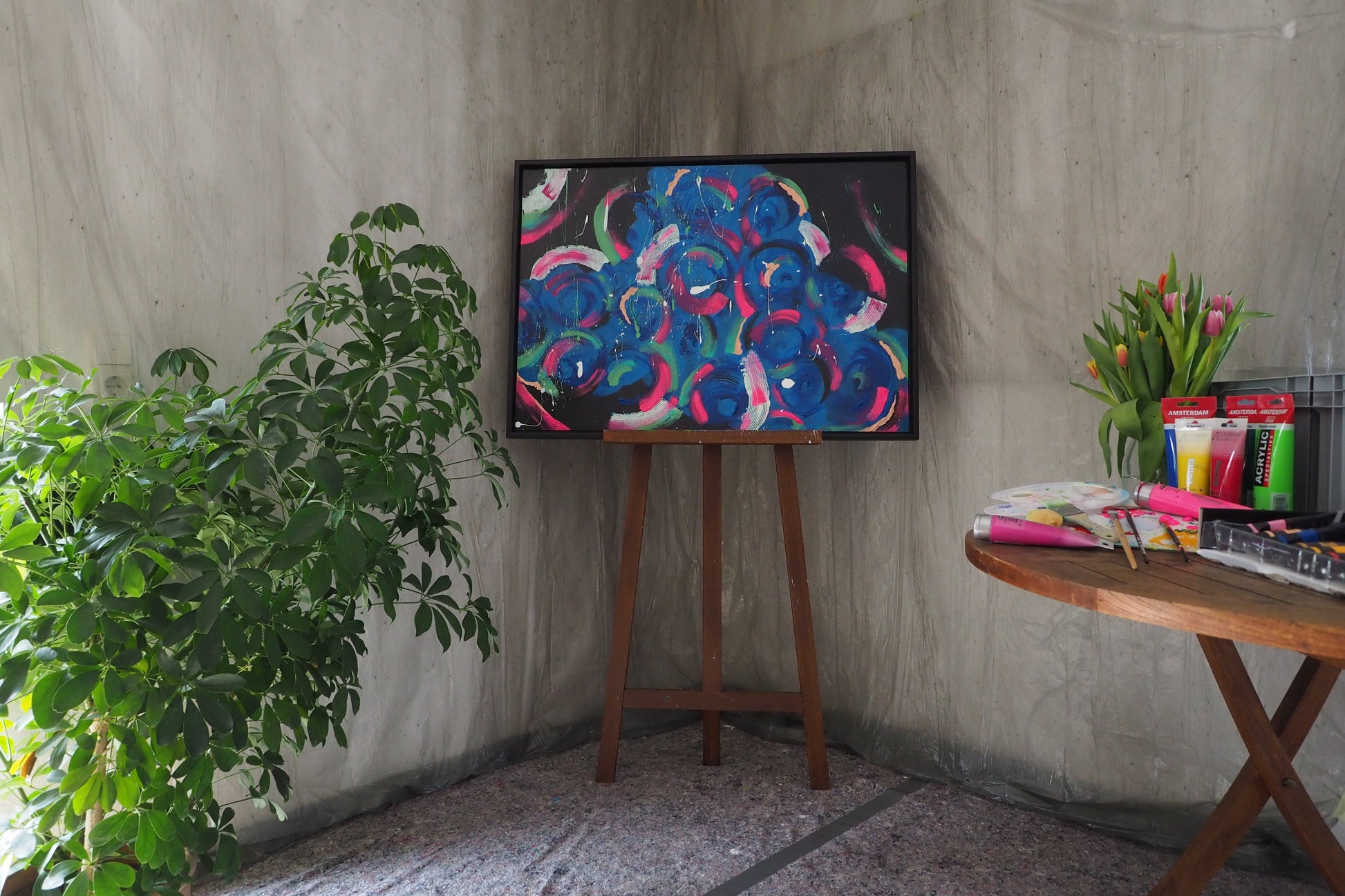 Das originale Kunstwerk „Tanzlust“ von Maridy Art, ein farbenfrohes abstraktes Gemälde auf einer Staffelei, steht neben einer grünen Pflanze. Ein runder Tisch mit Malutensilien und Tulpen runden die Szene vor Betonwänden ab.